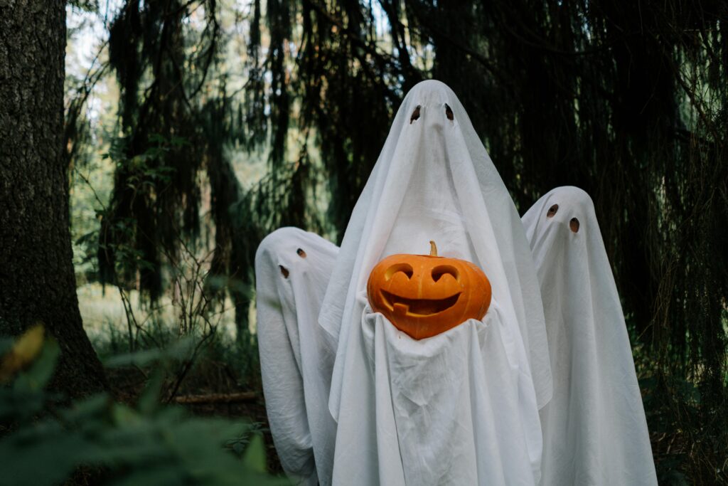 Three ghostly figures holding a carved pumpkin in a mysterious forest setting, perfect for Halloween imagery.