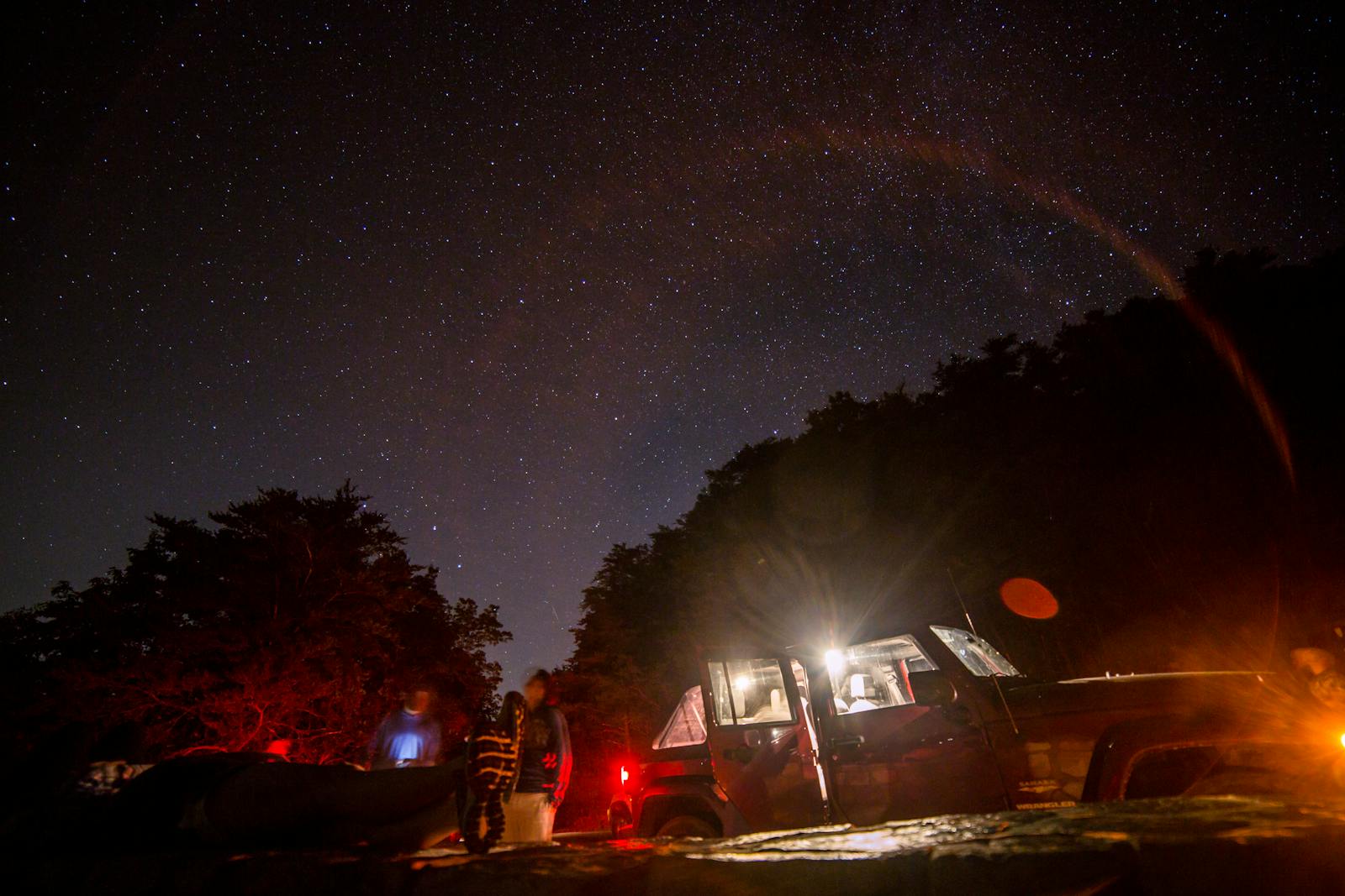 Starry night camping scene with a vehicle and blurred people illuminated by car lights in the wilderness.