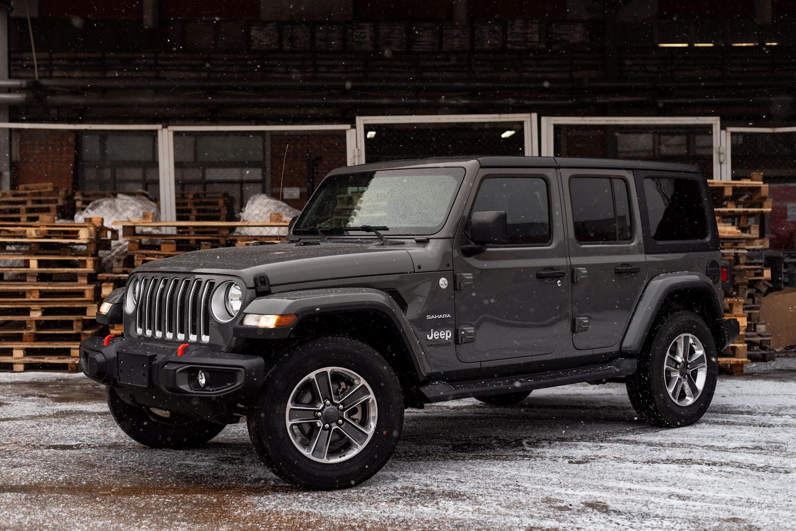 A Jeep Sahara parked in an industrial area with light snow, showcasing its rugged style and readiness for winter driving.